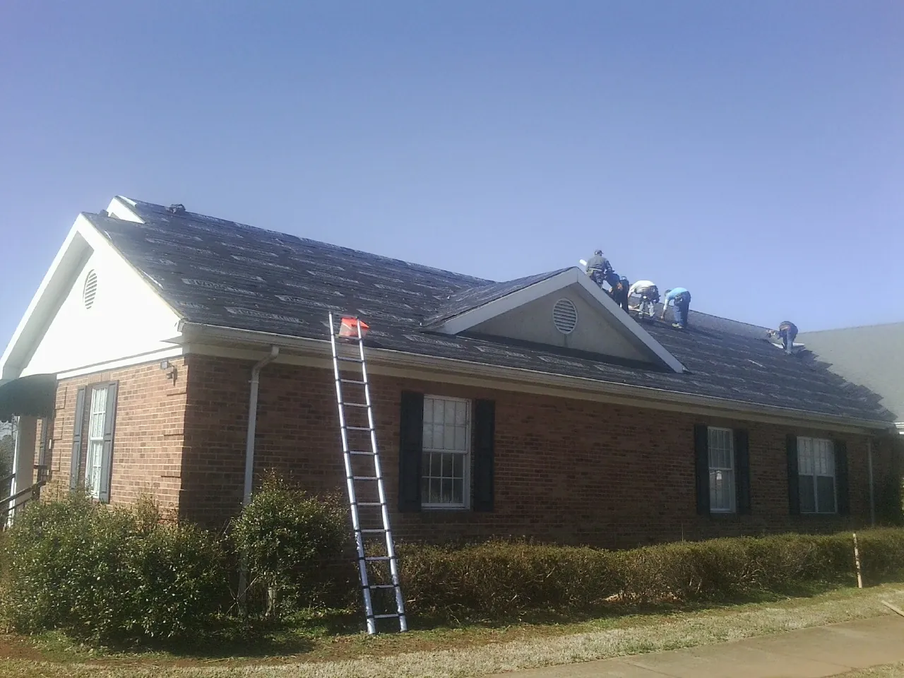 Crew tearing shingles off a brick ranch roof with a ladder set — Emergency Roof Repair work by our crew in Grimsley