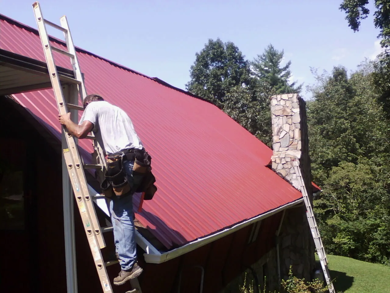 Roofer on a ladder beside a red metal roof and stone chimney — Soffit & Fascia Repair by Roofing Brothers in Grimsley